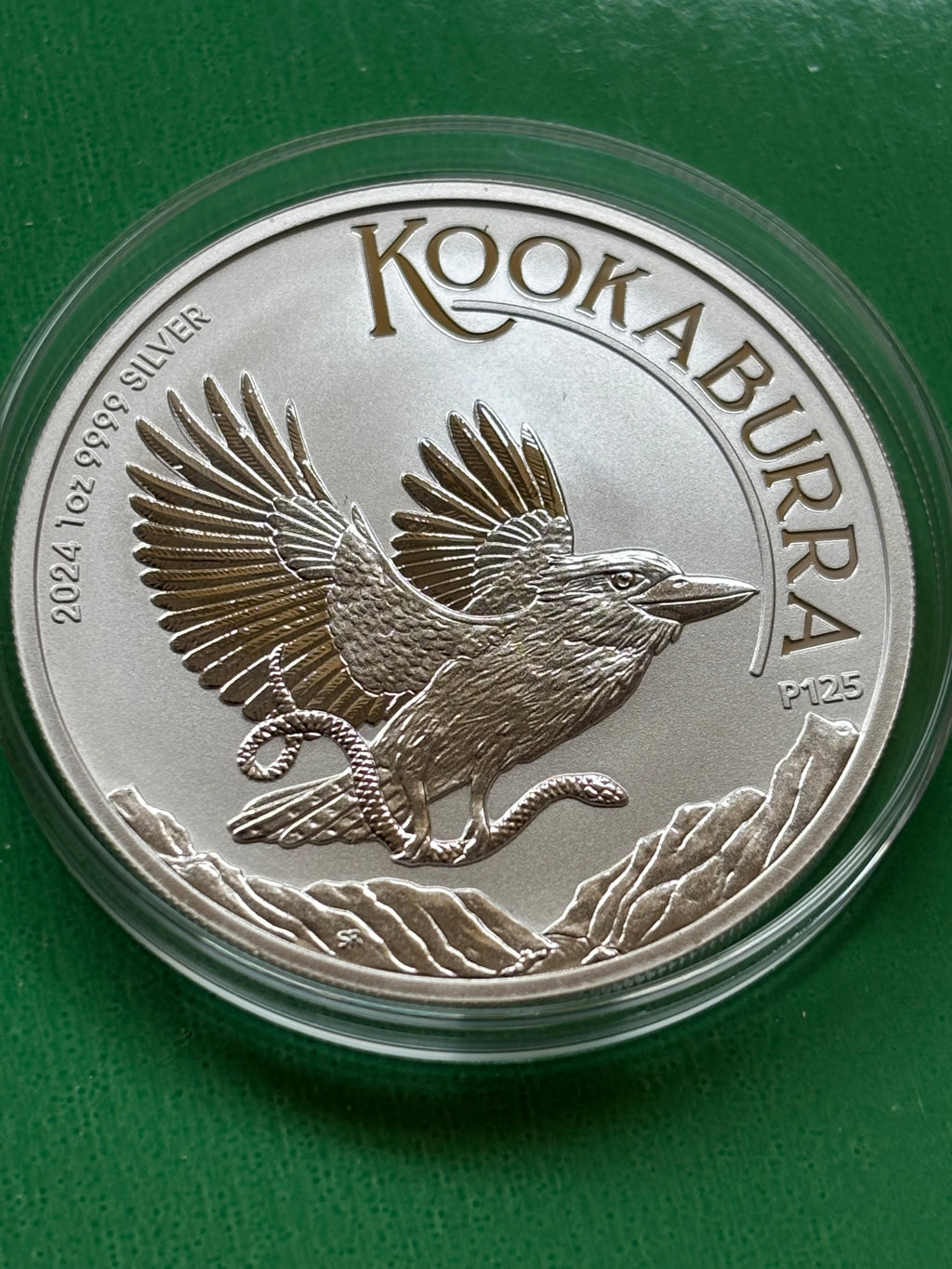 A 1oz .9999 Silver Kookaburra 2024 coin shows a kookaburra in flight holding a snake, surrounded by mountains, labeled "Kookaburra," with "2024," "P125," and purity details on a green background.