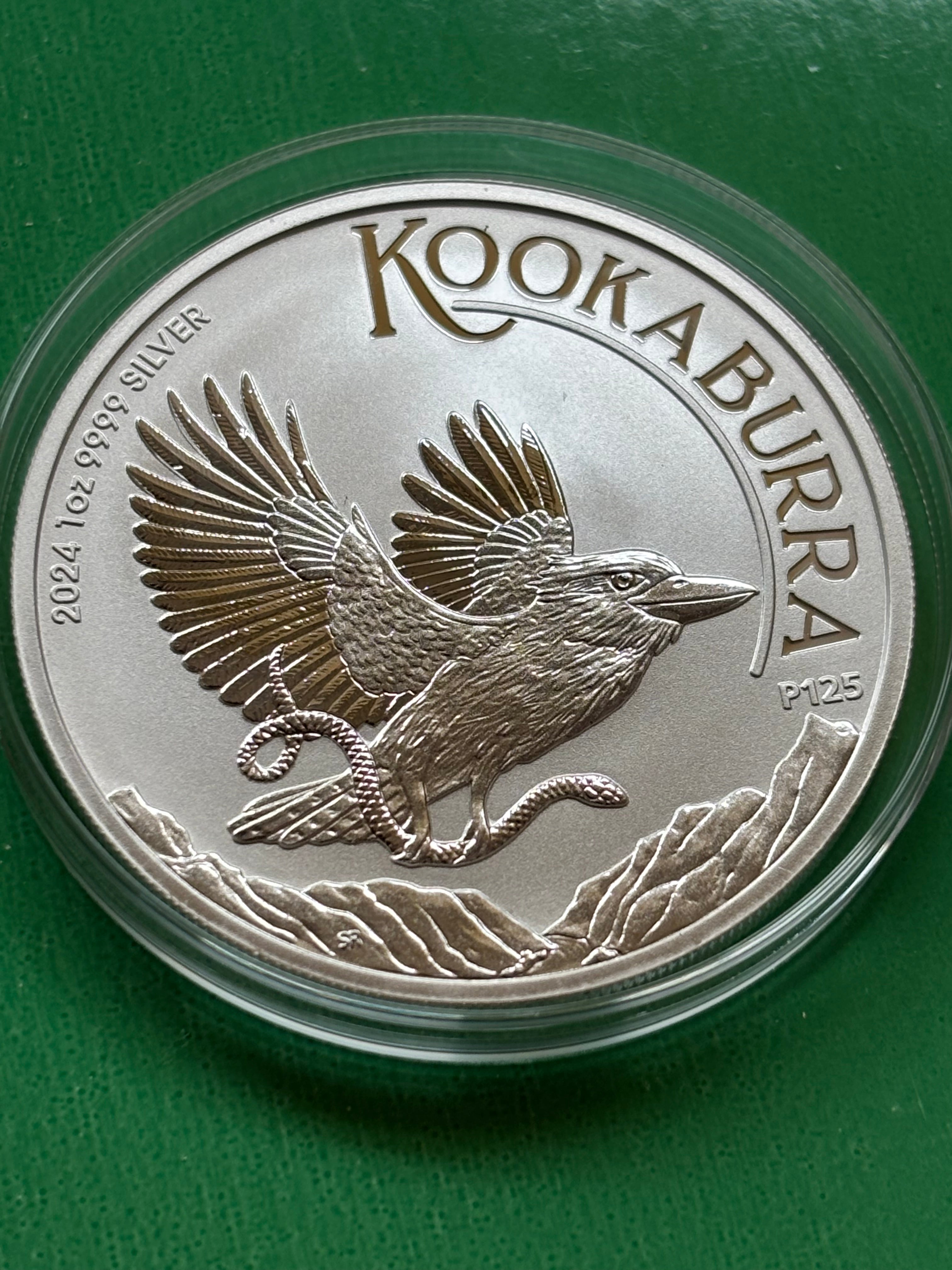 A 1oz .9999 Silver Kookaburra 2024 coin shows a kookaburra in flight holding a snake, surrounded by mountains, labeled "Kookaburra," with "2024," "P125," and purity details on a green background.