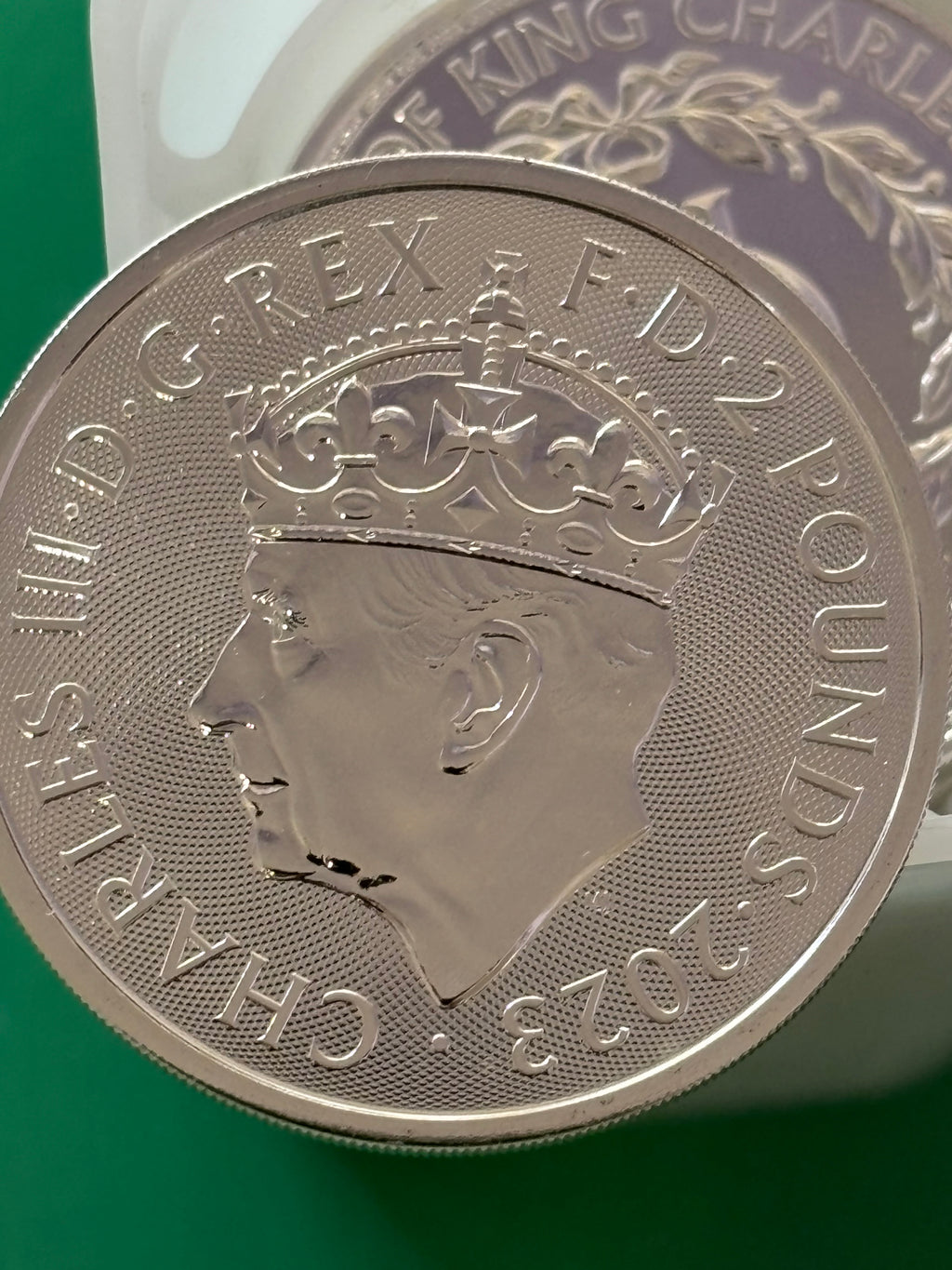 A close-up of a 1oz .999 Silver Coin Charles III Coronation, showing King Charles III in profile with inscriptions along the edge, set against a green background.