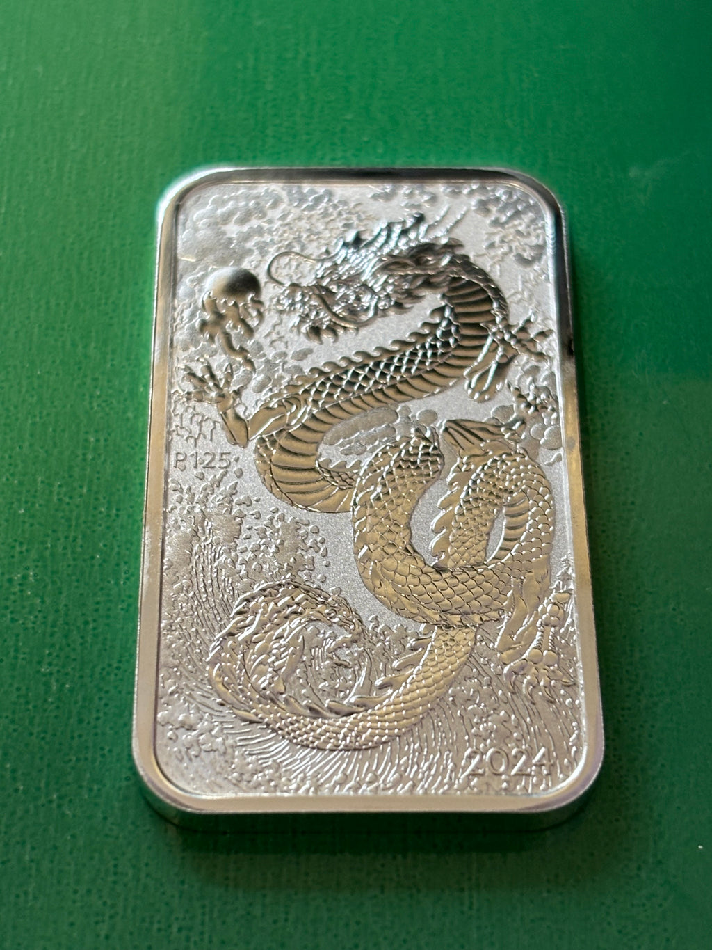 The 1oz .9999 Silver Bar Perth Mint Dragon 2024 displays an intricate engraved dragon with scales and whiskers on a textured background, marked with "2024" and "P125," and is shown resting on a green surface.
