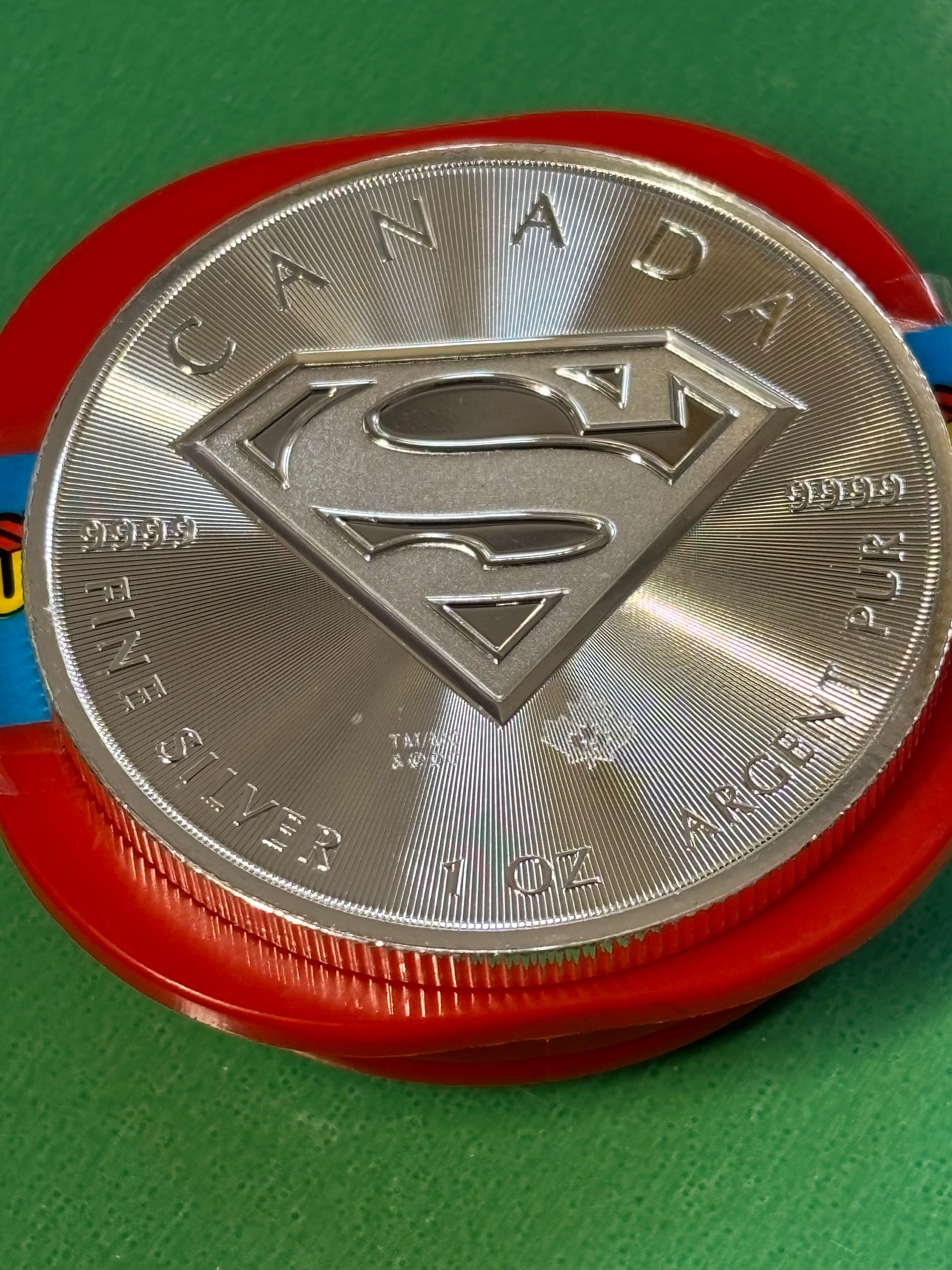 A 1oz .9999 Silver Coin Superman from Canada, featuring the Superman S logo and inscribed with "FINE SILVER 1 OZ ARGENT PUR" and "CANADA," sits on a red container against a green background.