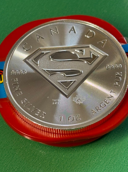 A 1oz .9999 Silver Coin Superman from Canada, featuring the Superman S logo and inscribed with "FINE SILVER 1 OZ ARGENT PUR" and "CANADA," sits on a red container against a green background.