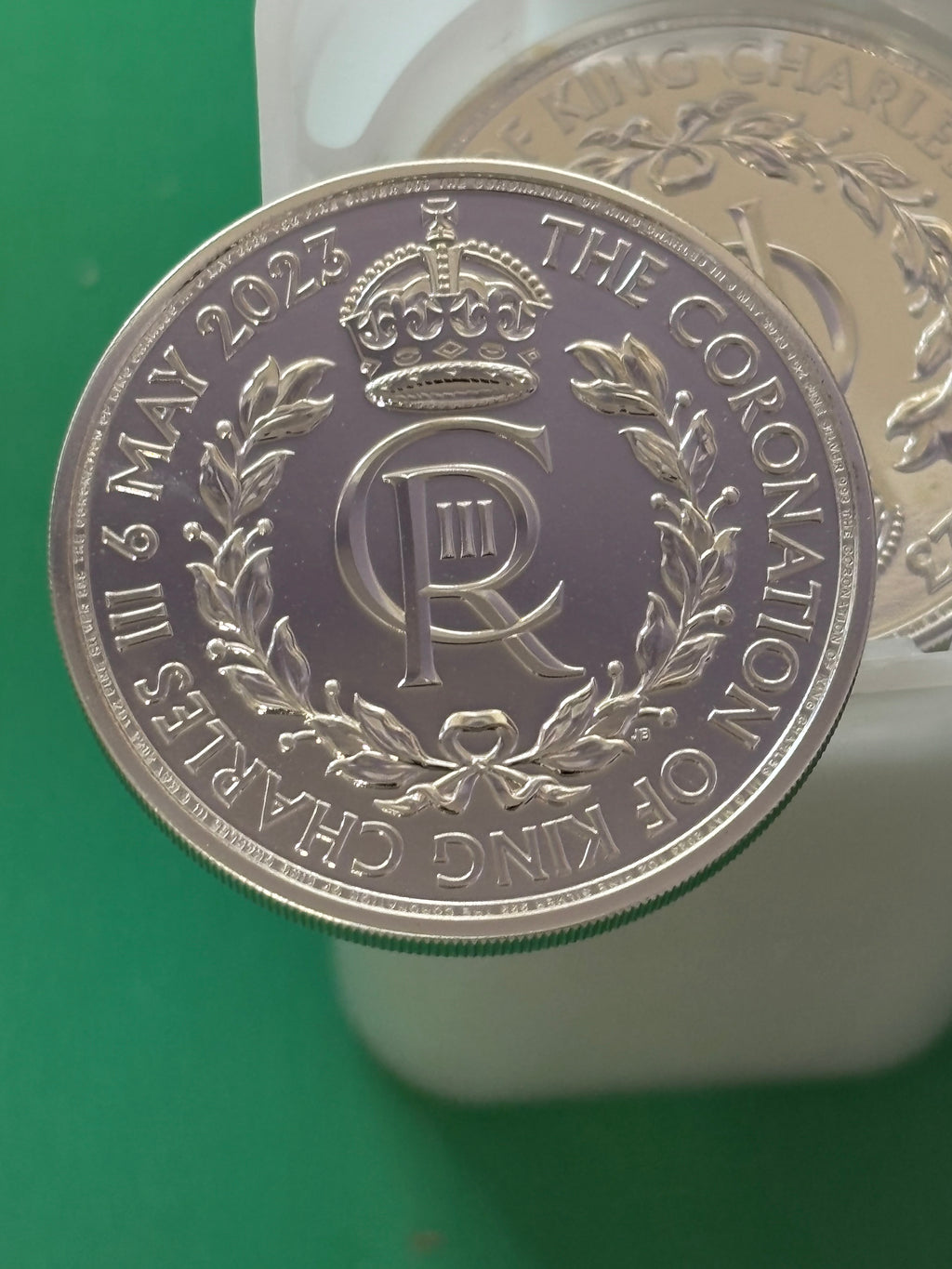 A close-up of the 1oz .999 Silver Coin Charles III Coronation shows a crown, "CR III," and a laurel wreath. The coin is inscribed with "THE CORONATION OF KING CHARLES III 6 MAY 2023" and held above a green surface.