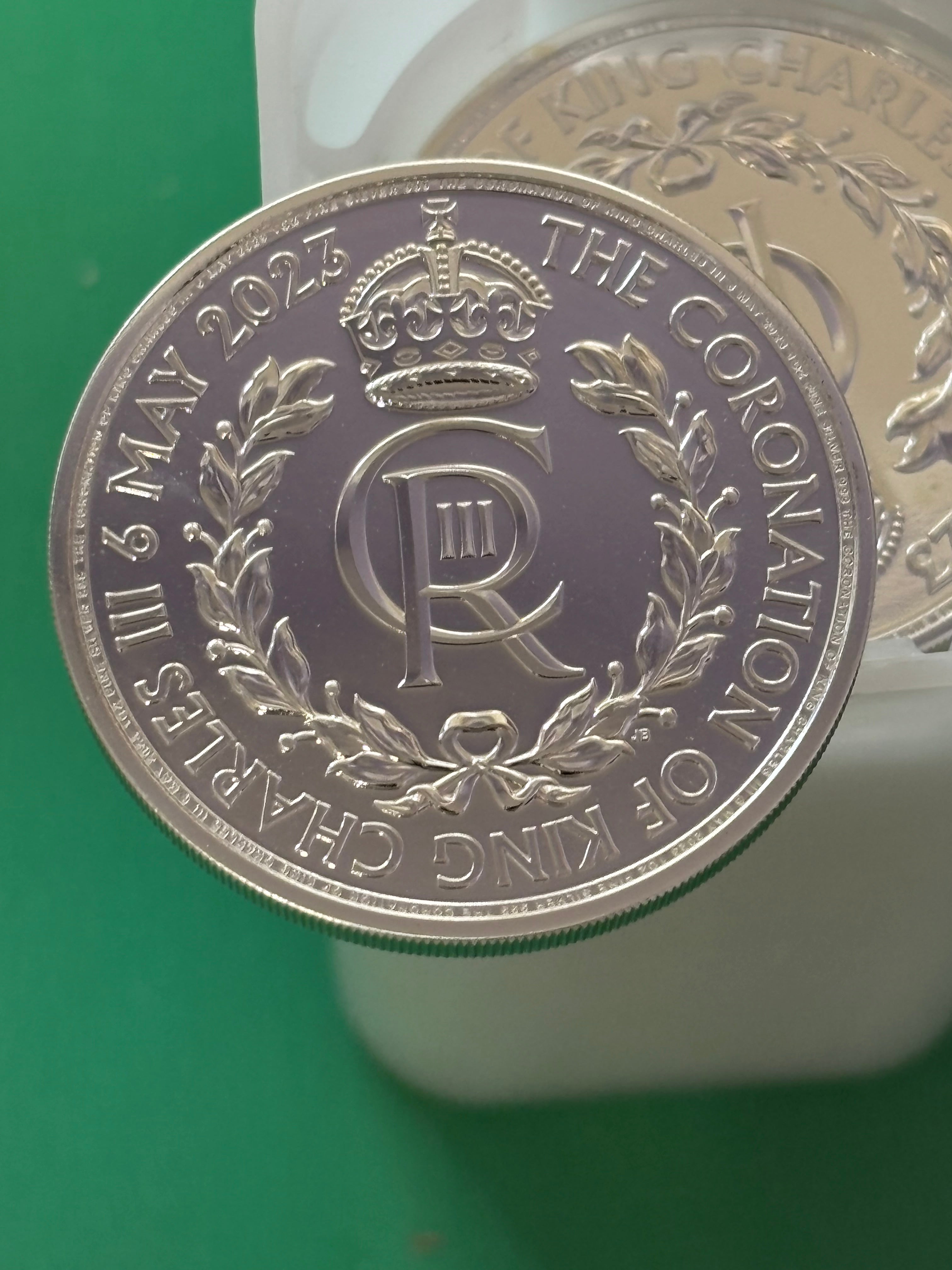 A close-up of the 1oz .999 Silver Coin Charles III Coronation shows a crown, "CR III," and a laurel wreath. The coin is inscribed with "THE CORONATION OF KING CHARLES III 6 MAY 2023" and held above a green surface.